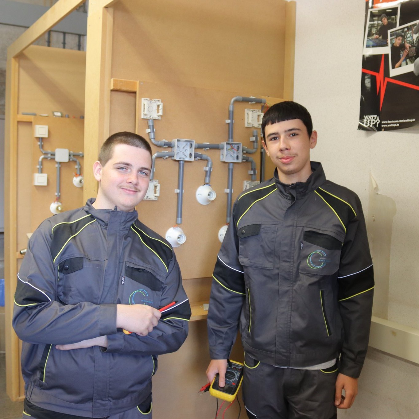 Deux élèves en tenue de travail devant leur panneau d’installation électrique